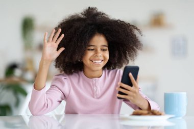 Neşeli Afro-Amerikalı kız, gür saçlı, video başında oturuyor, masada oturuyor, kurabiye yiyor, meyve suyu içiyor ya da kakao içiyor, cep telefonunu tutuyor, alet ekranına el sallıyor ve gülümsüyor, boşluğu kopyalıyor.