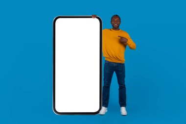 Pozitif stil, orta yaşlı siyah adam beyaz ekranlı ve gülümseyen büyük bir cep telefonunu işaret ediyor. Heyecan verici bir teklif sunuyor. Mavi stüdyo arka planı, model, tam boy.