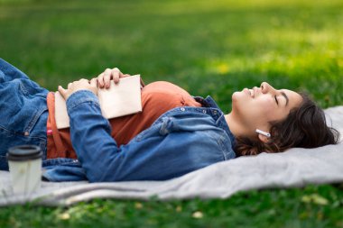 Dışarıda. Rahat ol. Ortadoğulu güzel bir kadın, parkta elinde kitapla uzanmış, gülümseyen bir Arap kadın, ekoseli gözleriyle dinleniyor ve kablosuz kulaklıkla müzik dinliyor.