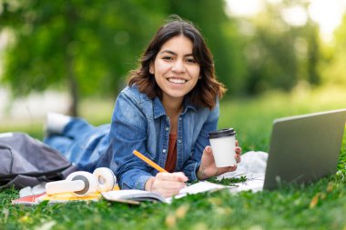 Şehir parkında dizüstü bilgisayarla ve not defteriyle çalışan güzel bir Arap kadını, çimlerin üzerinde uzanan ve kahve içen Orta Doğu 'lu bir kız öğrenci, dışarıda sınava hazırlanmanın keyfini çıkartıyor.