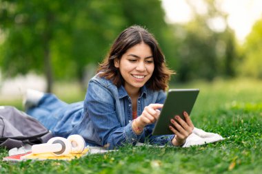 Gülümseyen genç Arap kadın, şehir parkında rahatlarken dijital tablette sosyal medyada geziniyor, mutlu bir ortadoğu öğrencisi çimlerde uzanıyor ve modern aletler kullanıyor, kopyalama alanı kullanıyor.