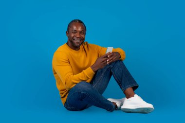 Rahat Afro-Amerikan orta yaşlı bir adam mavi stüdyo arka planında yerde oturuyor, yeni cep telefonu kullanıyor ve gülümsüyor, en yeni mobil uygulamaları kontrol ediyor,