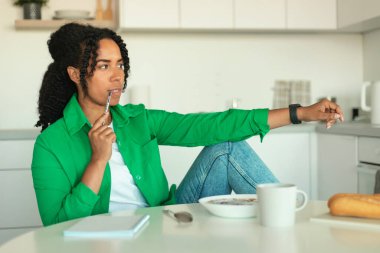 Düşünceli Afrikalı Amerikalı kadın, modern mutfakta oturmuş kahvaltı hazırlarken not alıyor ve diyet menülerini yiyor. Sağlıklı Beslenme ve Beslenme Konsepti