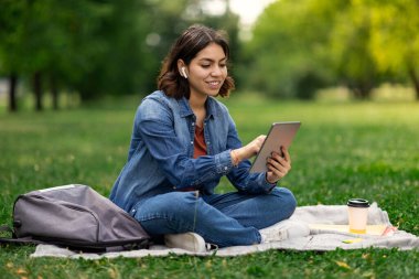 Gülümseyen Arap kız öğrenci parkta dinlenirken dijital tablet üzerinde internete göz gezdirirken, çimlerin üzerinde oturan mutlu Orta Doğulu kadın, Modern Gadget 'ı çalışma ya da boş zaman için kullanıyor,