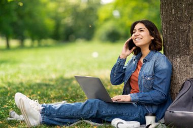 Gülümseyen Arap genç kız öğrenci cep telefonuyla konuşuyor ve şehir parkında ağacın altında otururken dizüstü bilgisayar kullanıyor. Mutlu Orta Doğulu kadın keyifli bir telefon görüşmesi yapıyor.