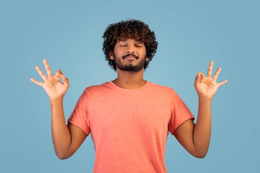 Pembe tişörtlü barışçıl kıvırcık Hintli adam zen jesti içinde el ele tutuşuyor ve gözlerini kapalı tutuyor mavi stdio arka plan üzerinde meditasyon yapıyor, kopyalama alanı. Stres atma kavramı