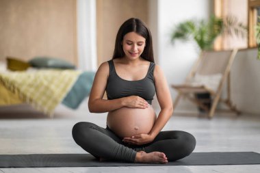 Doğum Konsepti. Yoga minderi üzerinde oturan ve evde meditasyon yaparken göbeğini kucaklayan aktif giyimde bekleyen güzel bir kadın göbeğine dokunuyor ve boşluğu kopyalıyor.
