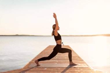 İnce, orta yaşlı bir kadın dışarıda yoga yapıyor, Savaşçı 1 poz veriyor, aktif kadın giysisini nehir kenarındaki ahşap rıhtımda eğitiyor, sabahları dışarıda meditasyon yapıyor, fotokopi alanı ile yan görüş yapıyor.