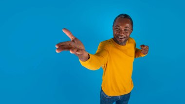 Neşeli orta yaşlı, günlük kıyafetli bir zenci elinde mavi stüdyo arka planında bir şey gösteriyor. Harika şeyler öneriyor. Reklam için fotokopi alanı olan bir panorama.