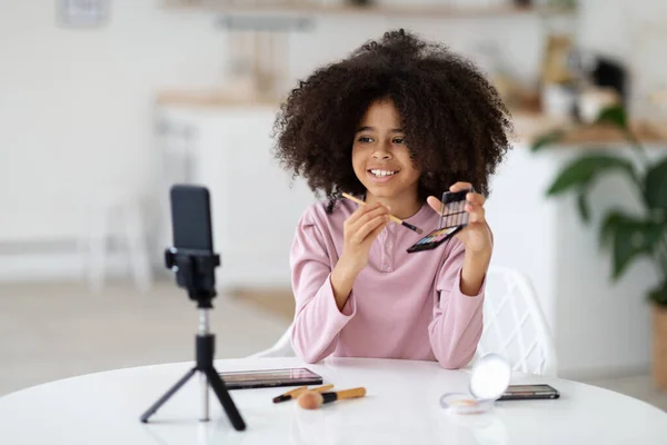Beauty blog for kids concept. Pretty cute black preteen girl recording video for her vlog, using smartphone set on tripod, child sitting at table at home, showing fancy cosmetics for little girls