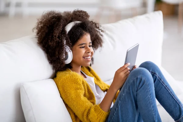 黒い子供の笑顔かわいいPreteen女の子はソファに座って ワイヤレスヘッドフォンと現代のデジタルタブレットを使用して インターネット上の漫画を見て 家のインテリア サイドビュー コピースペース — ストック写真
