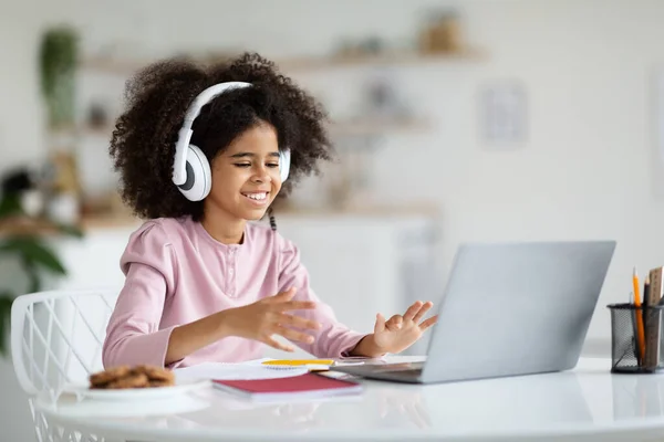 陽気なかわいいですアフリカ系アメリカ人の女の子とともにビデオ通話を持っています家庭でラップトップの前にテーブルに座って 無線ヘッドセットを使用して コンピュータ画面を見て ジェスチャー — ストック写真