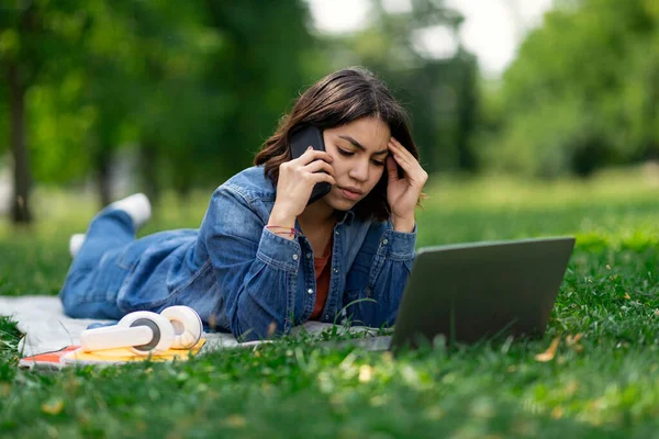 携帯電話で話している若いアラブ人女性を心配し 芝生の屋外に横たわっている間にノートパソコンを使用して 公園でコンピュータを勉強しながら 電話を持っている欲求不満中東の女性学生 フリースペース — ストック写真