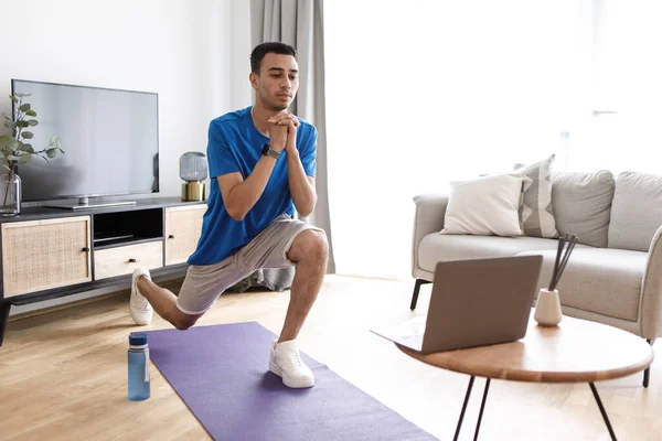 Young arab guy doing leg exercises on yoga mat in living room interior, watching online workout on laptop. Online class, new normal and body care at home