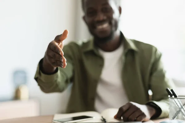 Partnership concept. Positive black man extending hand for handshake at camera, freelancer guy sitting at desk, greeting somebody and offering cooperation