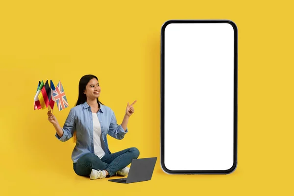 满意的年轻日本女学生 有许多不同的旗帜指向巨大的智能手机与空屏幕 孤立在黄色背景 学习语言 做广告和提供服务的应用程序 — 图库照片