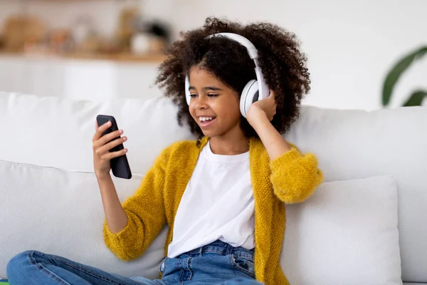 幸せなアフリカ系アメリカ人のPreteen女の子とともに巨乳毛でソファに座って自宅で 現代の無線ステレオヘッドフォンに触れ スマートフォンの画面を見て 音楽をオンラインで聴き スペースをコピー — ストック写真