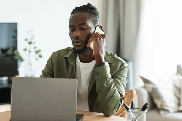 Serious african american male freelancer talking on cellphone and looking at laptop computer, working online from home. Business communication and distance job concept