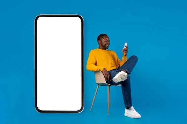 快乐的中年非洲男人 坐在椅子上 坐在巨大的智能手机旁 白色的空屏幕 在蓝色的工作室背景上使用手机 复制空间 — 图库照片