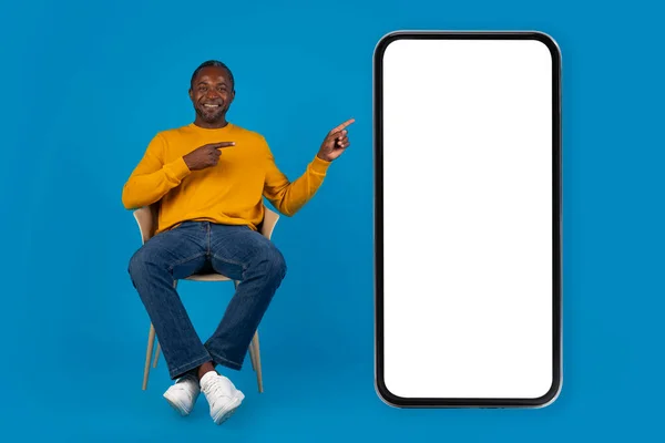 积极的中年黑人男子 穿着休闲装坐在椅子上 坐在巨大的智能手机旁 屏幕空白 神采奕奕 面带微笑 摆出一副蓝色画室的样子 装模作样 全身上下都是 — 图库照片