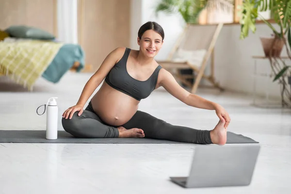 家庭运动 在客厅里用笔记本电脑对孕妇进行培训 让漂亮的准妈妈坐在瑜伽垫上 伸展腿肌肉 在线观看普拉提视频辅导 自由自在 — 图库照片