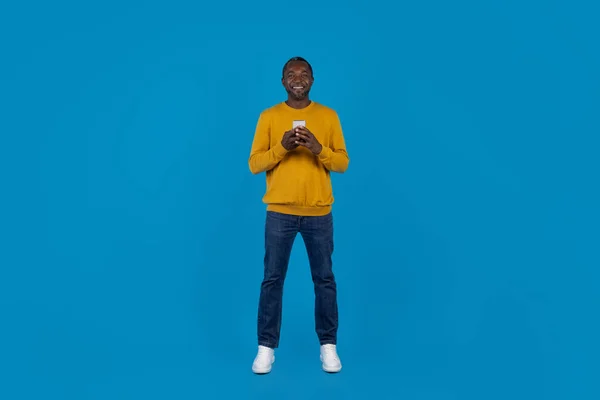 移动应用的概念 快乐英俊的非洲男子 身穿时髦休闲装 使用现代手机 带着蓝色工作室背景的微笑 全长镜头 全景与复制空间 — 图库照片