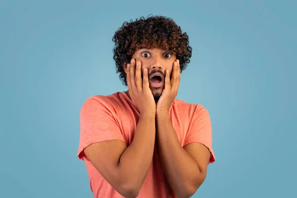 Frighten curly, bearded millennial eastern guy with open mouth touching his face and screaming, blue studio background, copy space, panorama. Human emotions and gestures concept