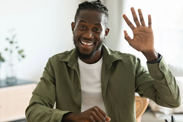 Greeting concept. Friendly black man waving at camera and smiling, positive guy gesturing hello, saying hi while having video call at home