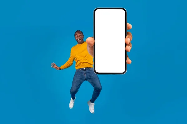 穿着时髦休闲装 情绪愉悦 英俊的中年黑人男子跳上带有白屏的现代手机 在蓝色工作室的背景 复制空间上展示出漂亮的应用程序 — 图库照片