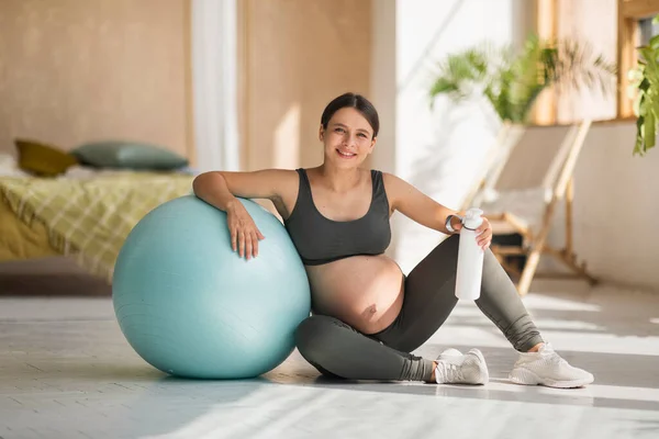 Exercise And Pregnancy. Beautiful Pregnant Woman In Activewear Sitting Near Big Fitball In Home Interior, Smiling Expectant Female Enjoying Domestic Trainings, Getting Ready For Childbirth