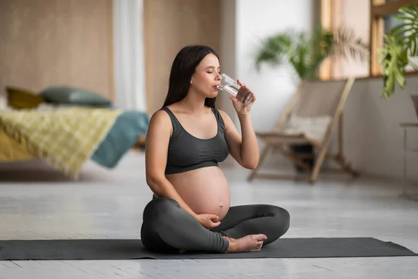 妊娠中の美しい女性が自宅でヨガを練習しながらガラスから水を飲む 妊娠中の腹を受け入れ 健康的なさわやかな飲み物を楽しんで スペースをコピーします — ストック写真