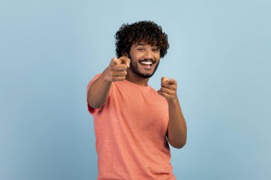 Hey sen! Jest konsepti. İki eliyle kamerayı gösteren çekici Hintli adam ve mavi stüdyo arka planında poz veren neşeli genç adam, fotokopi alanı.