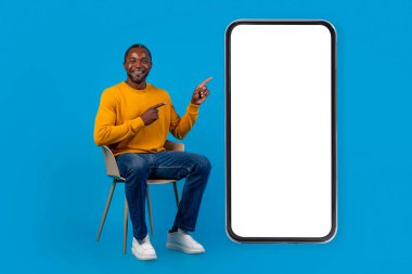 Neşeli orta yaşlı Afro-Amerikalı adam, gündelik sandalyede oturuyor ve beyaz ekranlı, gülümseyen, mavi stüdyo arka planında poz veren, uzun boylu akıllı telefonu işaret ediyor.