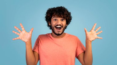 Şaşırmış kıvırcık Hintli adam mavi stüdyo arka planında el kaldırıyor ve çığlık atıyor, mutlu bin yıllık Hintli adam somurtuyor ve işaret ediyor, duygularını gösteriyor, kopyalama alanı.