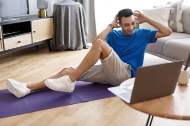 Genç Arap adam evde egzersiz yapıyor, çevrimiçi fitness videosu çekiyor ve karın kasları üzerinde çalışıyor, dizüstü bilgisayara bakıyor ve egzersiz yapıyor, uzayı kopyalıyor.