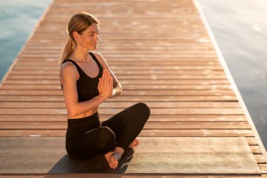 Kapalı Elleri ve Kapalı Gözleriyle Güzel Rahatlamış Kadın meditasyon yapıyor, Aktif Giyimde Gülümseyen Orta yaşlı Kadın Yoga yapıyor, Tahta İskele 'de Nehir' in yanında oturuyor, Uzay 'ı kopyalıyor.