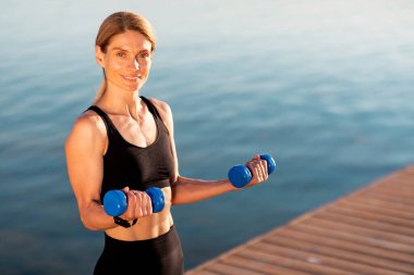 Gülümseyen Orta Çağ Kadın Sporcusu Dışarda Dambıllarla Egzersiz Yapıyor, Aktif Kadın Giyimi Portresi Nehir kenarındaki Ahşap İskele 'de, Formda Bayan Vücut Çalışmalarından Hoşlanıyor, Kopya Uzay