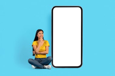 Sarı tişörtlü, dalgın gülümseyen Çinli kadın Lotus pozisyonunda oturur, boş ekranlı, mavi arka planda, stüdyoda izole edilmiş büyük akıllı telefona bakar. Çalışma fikri, satılık uygulama, reklam ve teklif