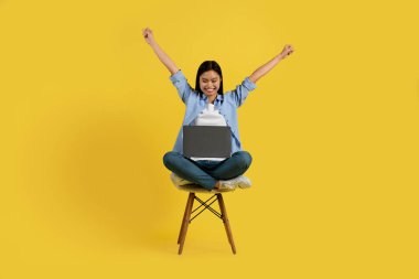Gülümseyen genç Çinli kız öğrenci dizüstü bilgisayarla sandalyede oturuyor, ellerini kaldırıyor, zafere seviniyor, sarı arka planda, stüdyoda izole edilmiş. Uzaktan ve dijital göçebe eğitimi, reklam ve teklif, yeni normal