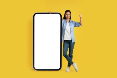 Sıradan gösteri yapan mutlu genç Çinli kız öğrenci, boş ekranlı, sarı arka planda, stüdyoda izole edilmiş büyük bir akıllı telefonun yanında duruyor. Eğitim ve çalışma için uygulama ve teknoloji, reklam ve sunum