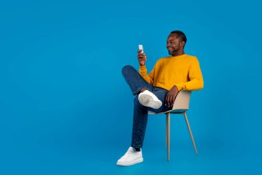 Rahat Afro-Amerikan orta yaşlı bir adam mavi stüdyo arka planında sandalyede oturuyor, yepyeni bir akıllı telefon kullanıyor ve gülümsüyor, kadınlarla sohbet ediyor, kopya alanlı panorama