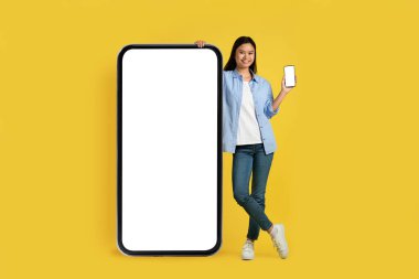 Yeni uygulama, dijital ve reklam teklifi. Sıradan gösteriler yapan gülümseyen genç Japon bayan öğrenci, sarı arka planda izole edilmiş, boş ekranlı, kocaman bir akıllı telefonun yanında duruyor. Çalışma ve alışveriş için araç