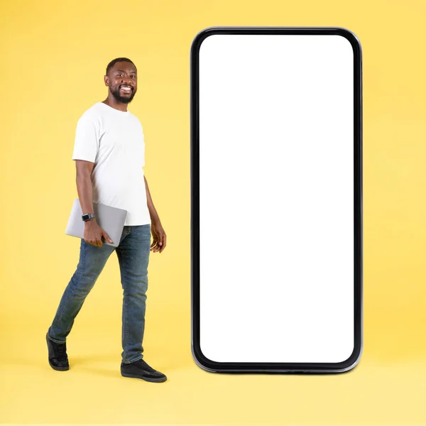 快乐的黑人男人靠近大型智能手机空白屏幕 手持笔记本电脑在黄色背景下行走在工作室里 移动应用程序广告 正方形 全长镜头 — 图库照片