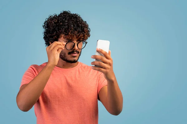 Shocked indian guy with curly hair in casual outfit checking newest mobile application or online offer, looking at smartphone screen, removing eyeglasses, blue studio background, copy space
