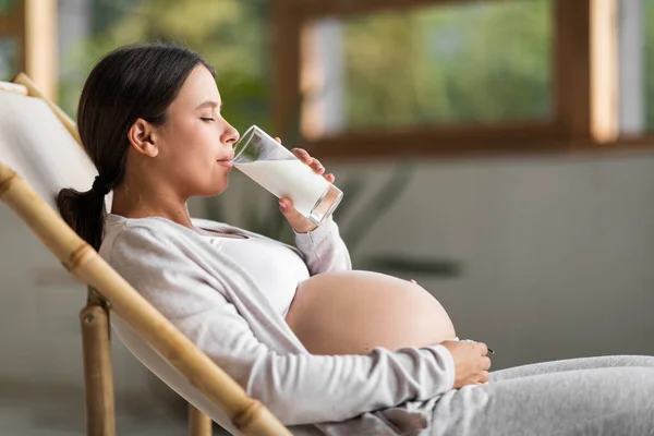 年轻美丽的怀孕妇女坐在椅子上 喝着玻璃杯中的牛奶 在家中享受健康钙饮料的迷人的千年妇女的侧视图 复制空间 — 图库照片