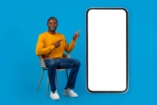 快乐的中年非洲裔美国男人 随意坐在椅子上 指着有白色空屏幕的巨大智能手机 微笑着 摆出一副蓝色画室的样子 装腔作势 全身上下都是 — 图库照片