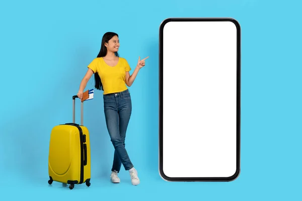 身穿黄色T恤 面带微笑的年轻韩国女士拿着护照 用手指着屏幕空的巨大智能手机 与蓝色背景隔离 工作室 在线地图 旅行应用程序 广告和提供服务 — 图库照片