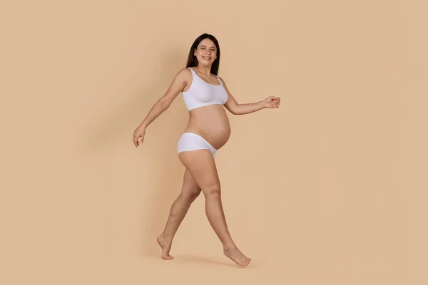 Pregnancy Concept. Cheerful Young Pregnant Woman In Underwear Walking Over Beige Background In Studio And Smiling At Camera, Full Length Shot Of Beautiful Expectant Woman Wearing Lingerie, Copy Space