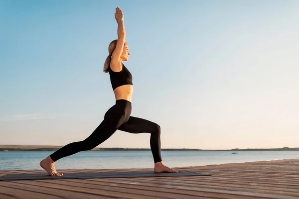 Athletic Middle Aged Woman Practicing Yoga On Wooden Pier Outdoors, Sporty Smiling Female In Activewear Enjoying Training Outside, Standing In Warrior 1 Pose, Side View With Copy Space