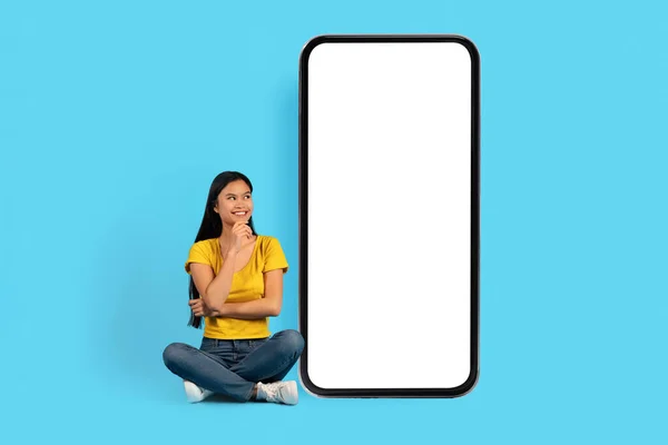 穿着黄色T恤的富有沉思的年轻中国女士坐在莲花的位置上 看着有着空白屏幕的巨大智能手机 蓝色背景 工作室 学习的想法 销售的应用程序 广告和提供 — 图库照片